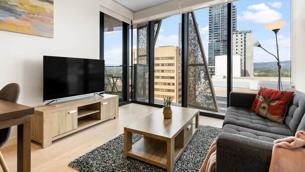 A modern living room with a flat-screen TV, wooden furniture, a grey sofa, and large windows offering a city view. - The 2BR City Escape - Stylish and Mordern (Adelaide)