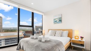 A modern hotel room with a large bed, a nightstand with a lamp, and a view of the cityscape through a large window. - The 2BR City Escape - Stylish and Mordern (Adelaide)