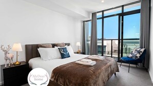A modern hotel room with a large bed, two bedside tables, a nightstand, a chair, and a view of the city through large windows.