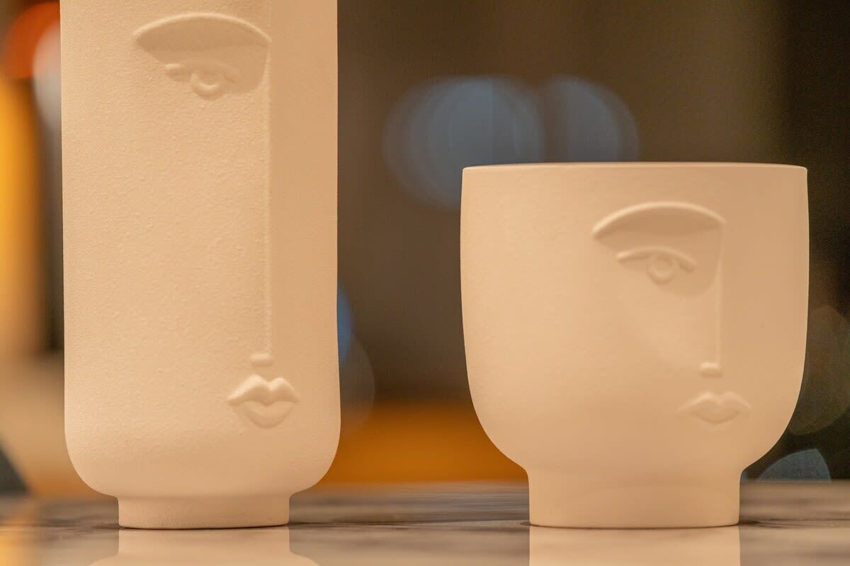 Twee witte keramische bekers met reliëfdecoraties, één met een gezicht en de andere met een neus en lippen.
