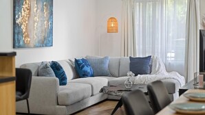 A modern living room with a grey sectional sofa, a small coffee table, and a dining table with chairs. There is a large abstract painting on the wall and a floor lamp near the window. - Hidden Gem of CBD - 3BR Apt-Free Parking-WiFi (Adelaide)