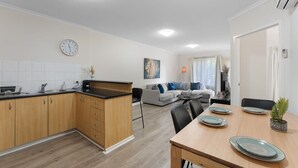 A modern kitchen with wooden cabinets, a dining table set with plates, and a living area with a sofa and a painting. - Hidden Gem of CBD - 3BR Apt-Free Parking-WiFi (Adelaide)