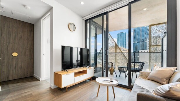 Un salón moderno con una televisión de pantalla plana, suelo de madera, un sofá y un amplio ventanal corredero que da a un balcón exterior con vistas a la ciudad.