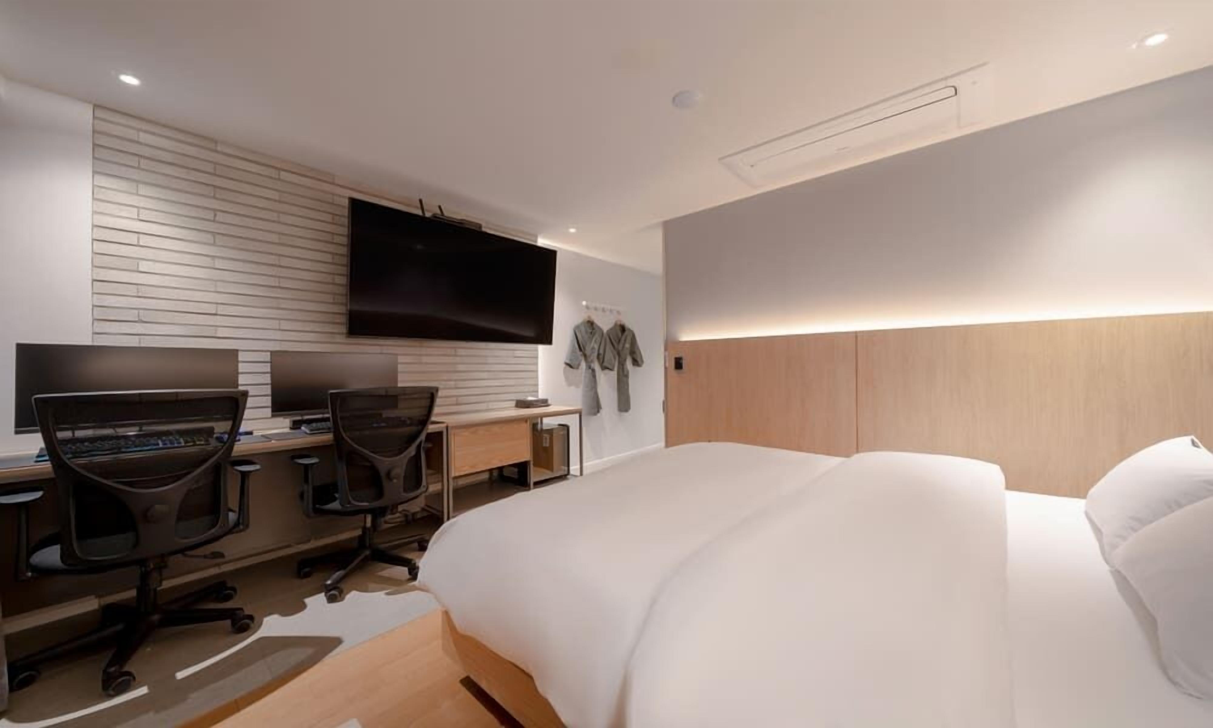 A modern hotel room with a large bed, a desk with two computers, a flat-screen TV, and a wooden headboard.