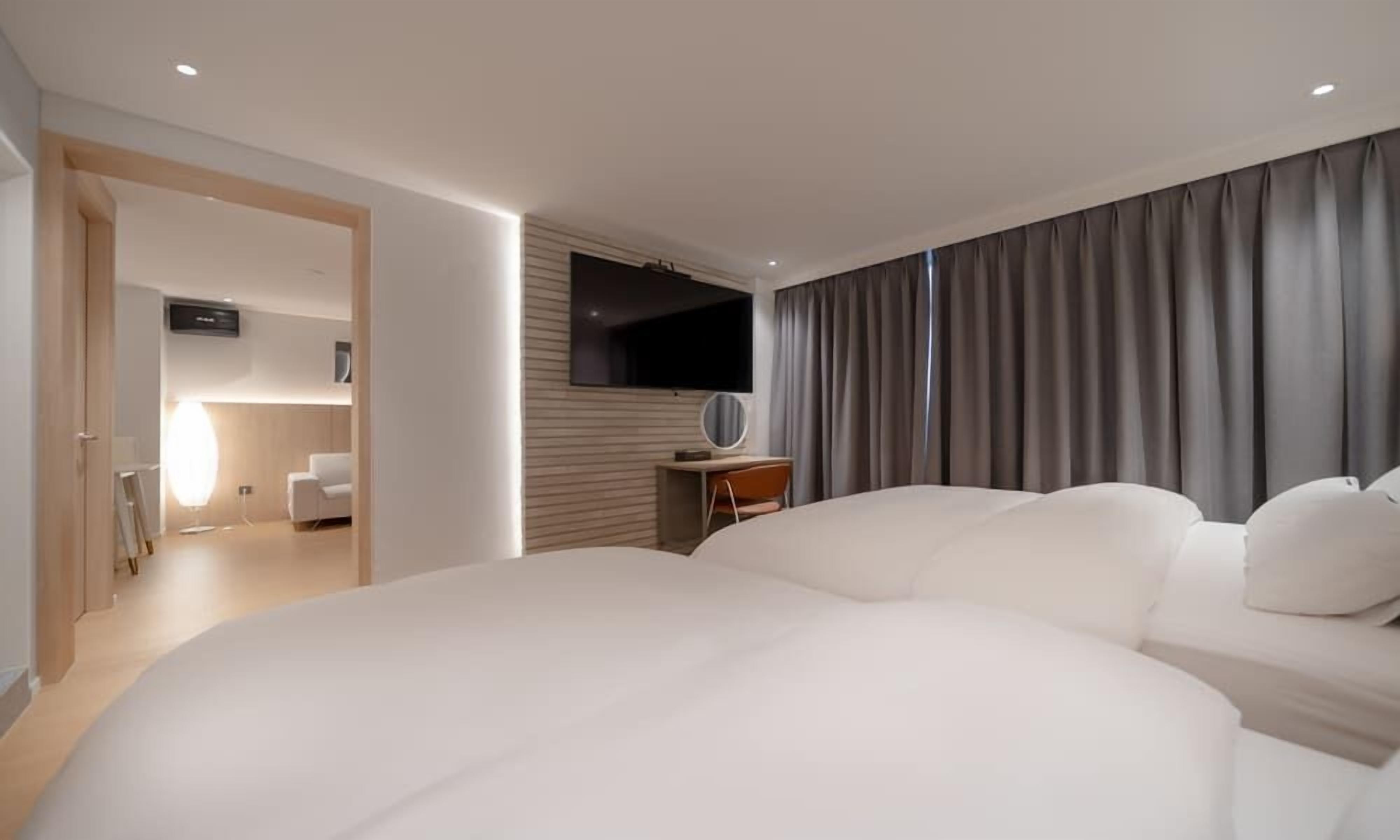 A modern hotel room with a large bed, a desk with a chair, a flat-screen TV, and a large window with curtains.