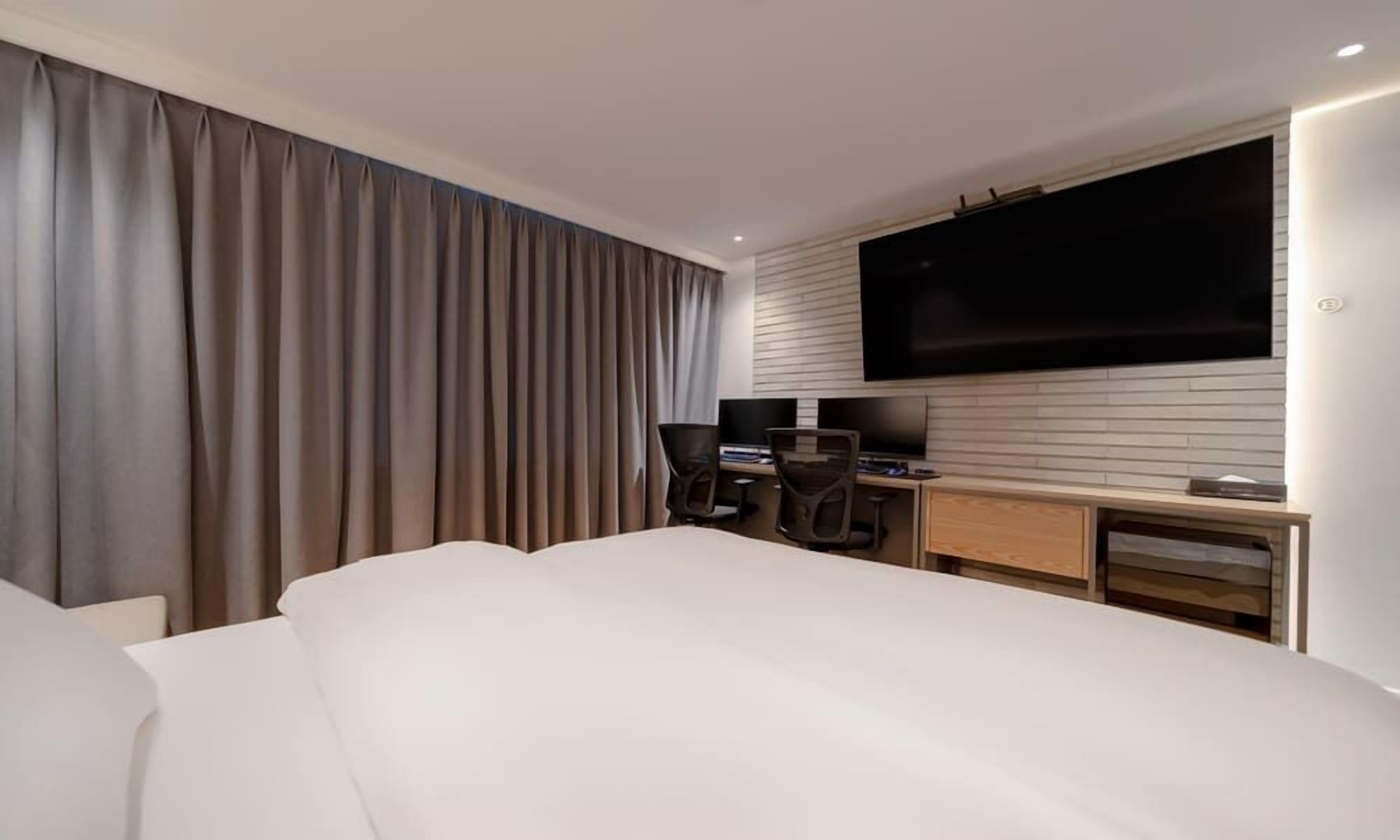 A modern hotel room with a large bed, a desk with a computer, a TV mounted on the wall, and a large window with curtains.