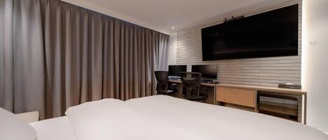 A modern hotel room with a large bed, a desk with a computer, a TV mounted on the wall, and a large window with curtains.