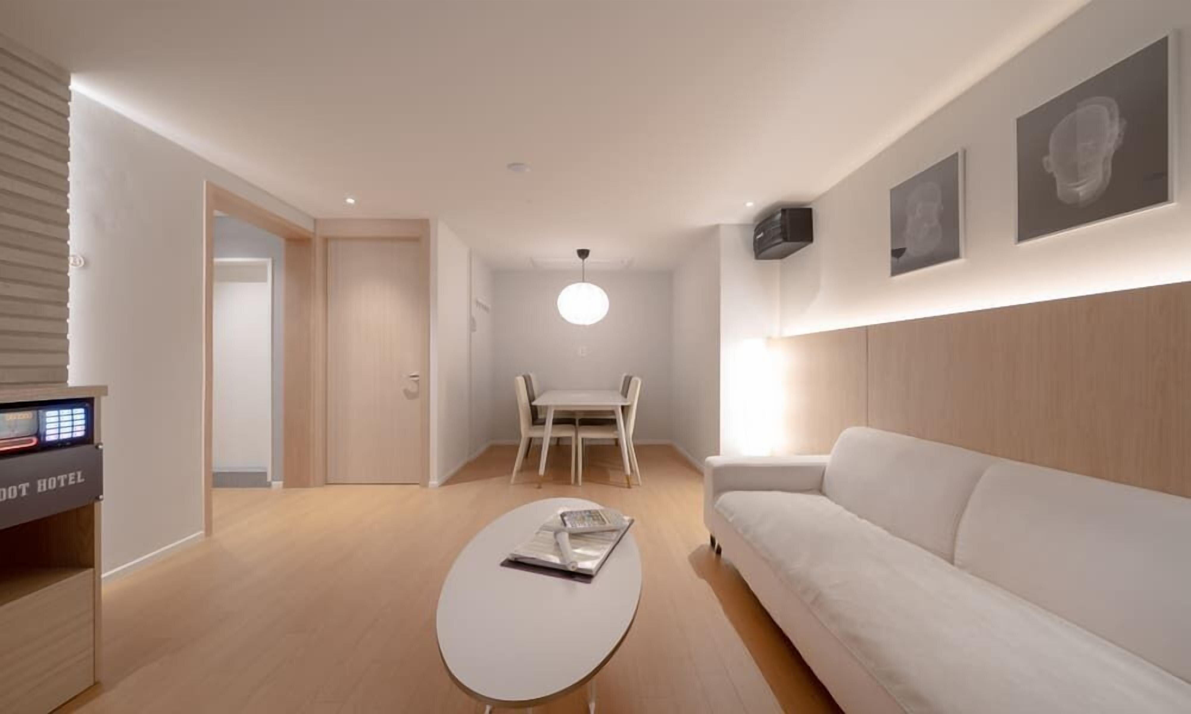 A modern hotel room with a white sofa, a dining table with chairs, a flat-screen TV, and a ceiling light.