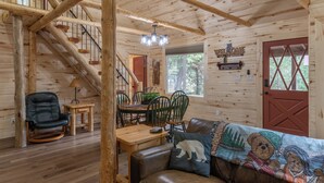 A wooden cabin interior with a dining area, a sofa with a bear-themed blanket, and a staircase.