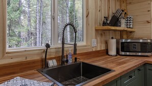 A kitchen with a wooden interior, a stainless steel sink, a microwave, and a window overlooking a forest.