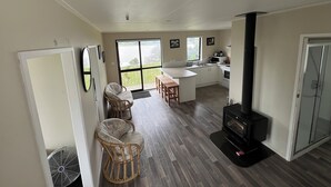 A modern kitchen with a central island, a dining area with chairs, a fireplace, and large windows offering a view. - Peaceful riverfront cottage in tiny northland town (Te Kopuru)