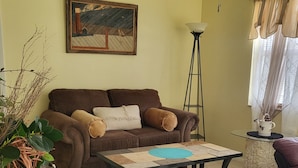 Ein Wohnzimmer mit braunem Sofa, einem Couchtisch, einem violetten Hocker, einem Bild an der Wand und einer Pflanze.
