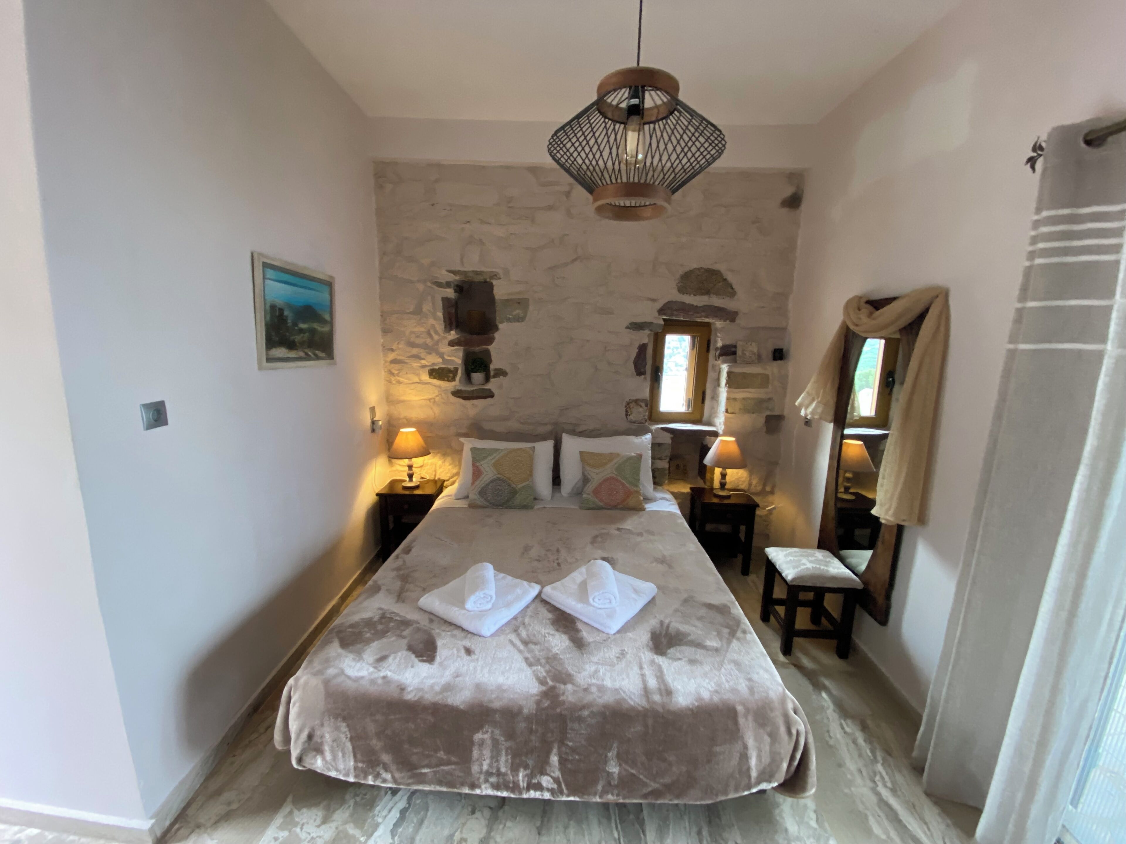 A bedroom with a stone wall, a bed with a beige cover, two bedside tables with lamps, a hanging light fixture, and a mirror on the wall.