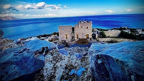 A stone building with a tower, overlooking a coastal area with a clear blue sea and distant mountains.