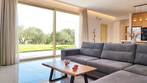 A modern living room with a grey sectional sofa, a wooden coffee table, and a large sliding glass door leading to a garden. - Fence off , surrounded by large olive grove 200m from the sandy beach (Ζάκυνθος)