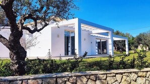 A modern white house with a stone wall in the foreground, surrounded by greenery and trees. - Fence off , surrounded by large olive grove 200m from the sandy beach (Ζάκυνθος)