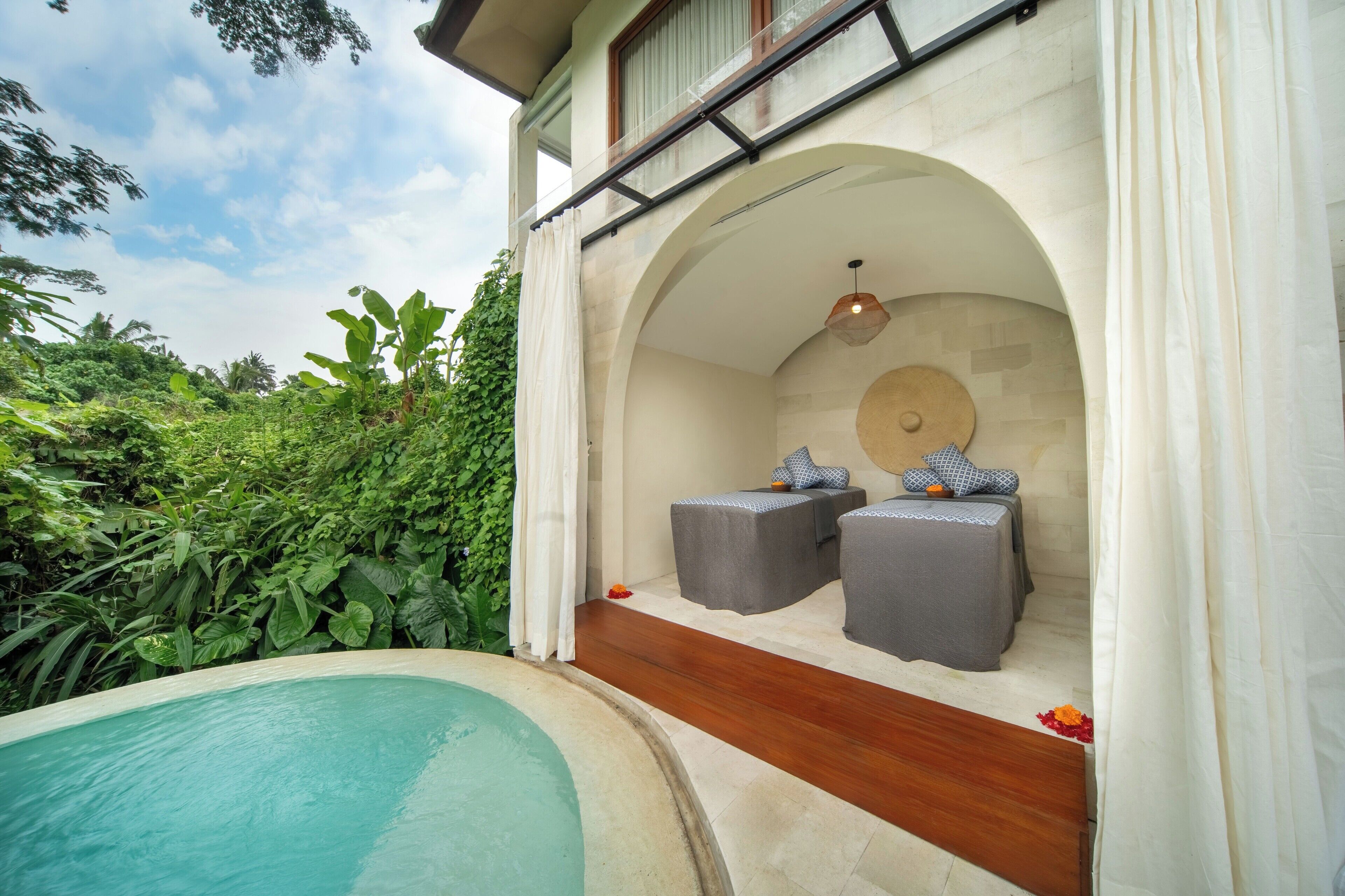 Un área de piscina privada con un deck de madera y dos zonas de estar acolchadas bajo un techo arqueado.