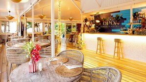 Un espace extérieur aménagé avec des chaises en osier et des tables rondes, un bar doté d’un comptoir en bois et un plafond orné de panneaux de bois disposés horizontalement.
