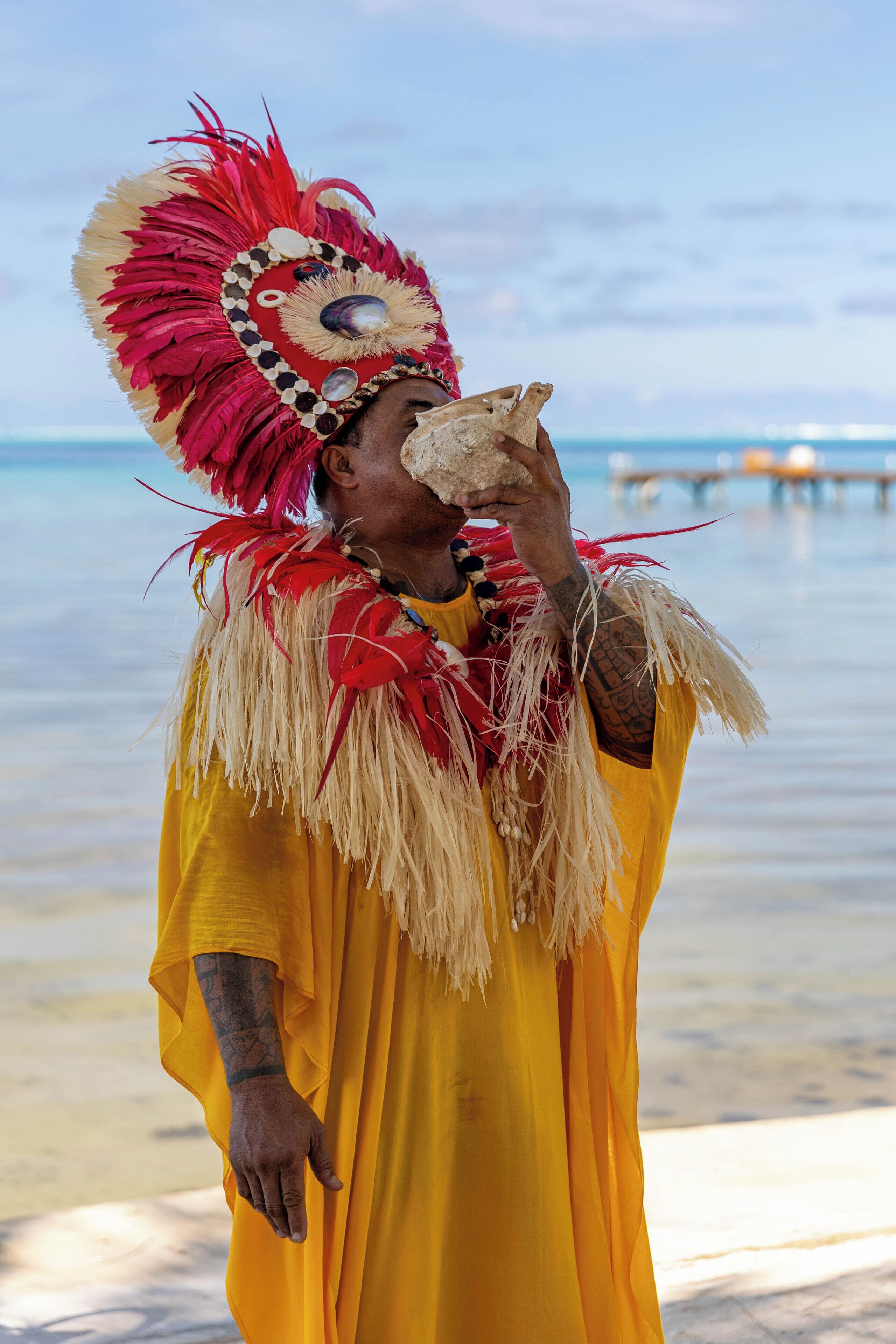 A person in a yellow garment and red headdress standing by a beach with a pier in the background.