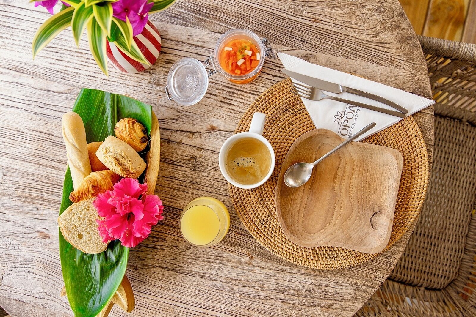 Ett träbord med en frukostuppsättning som inkluderar croissanter, bröd, en blomma, en kopp kaffe, ett glas juice och en liten burk med sylt.