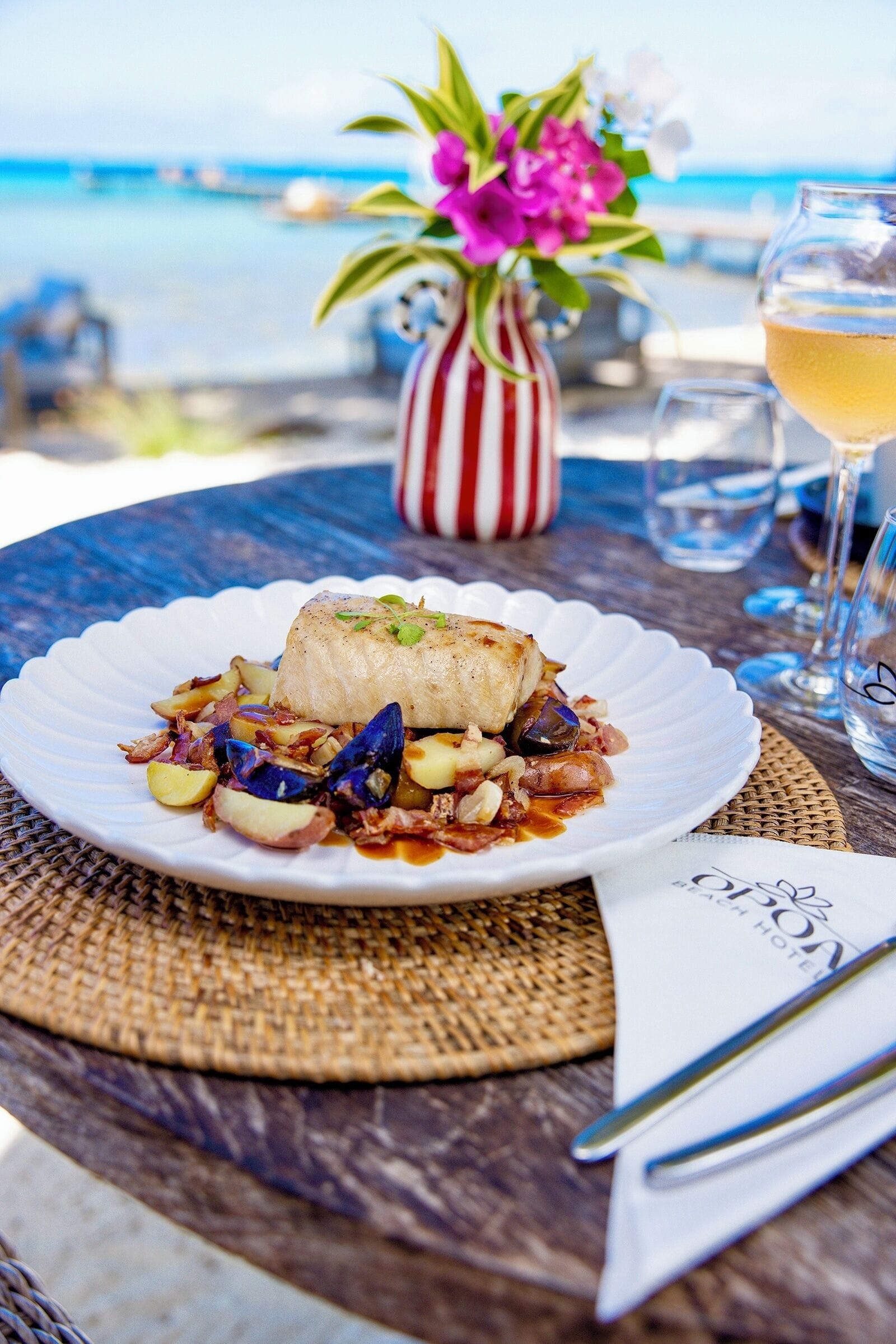 Prato de mariscos sobre um prato branco com um jogo americano de vime, um copo de vinho branco e um vaso listrado com flores em uma mesa com vista para o mar.