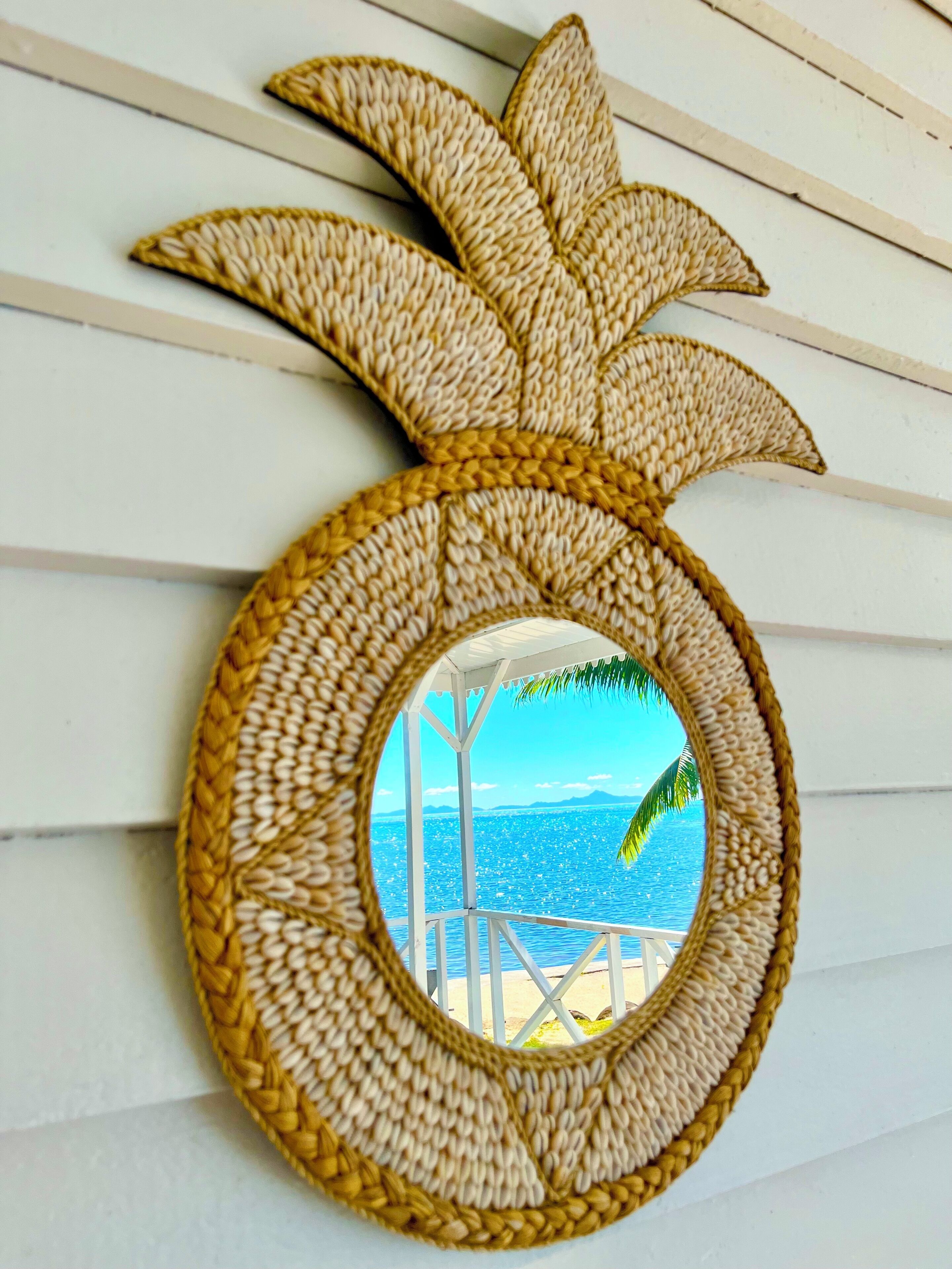 Un espejo de piña trenzada sobre una pared blanca, con vista al océano y palmeras.