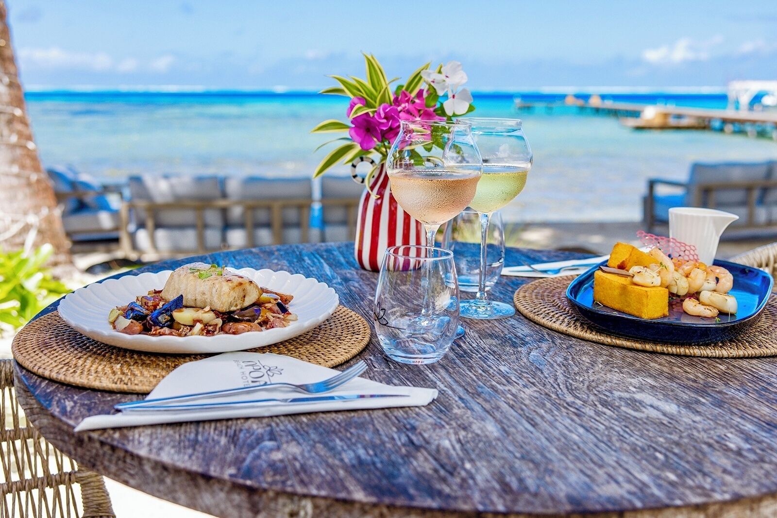 Una mesa servida con platos de mariscos, una jarra de bebida y un florero con flores, con vista a una playa.