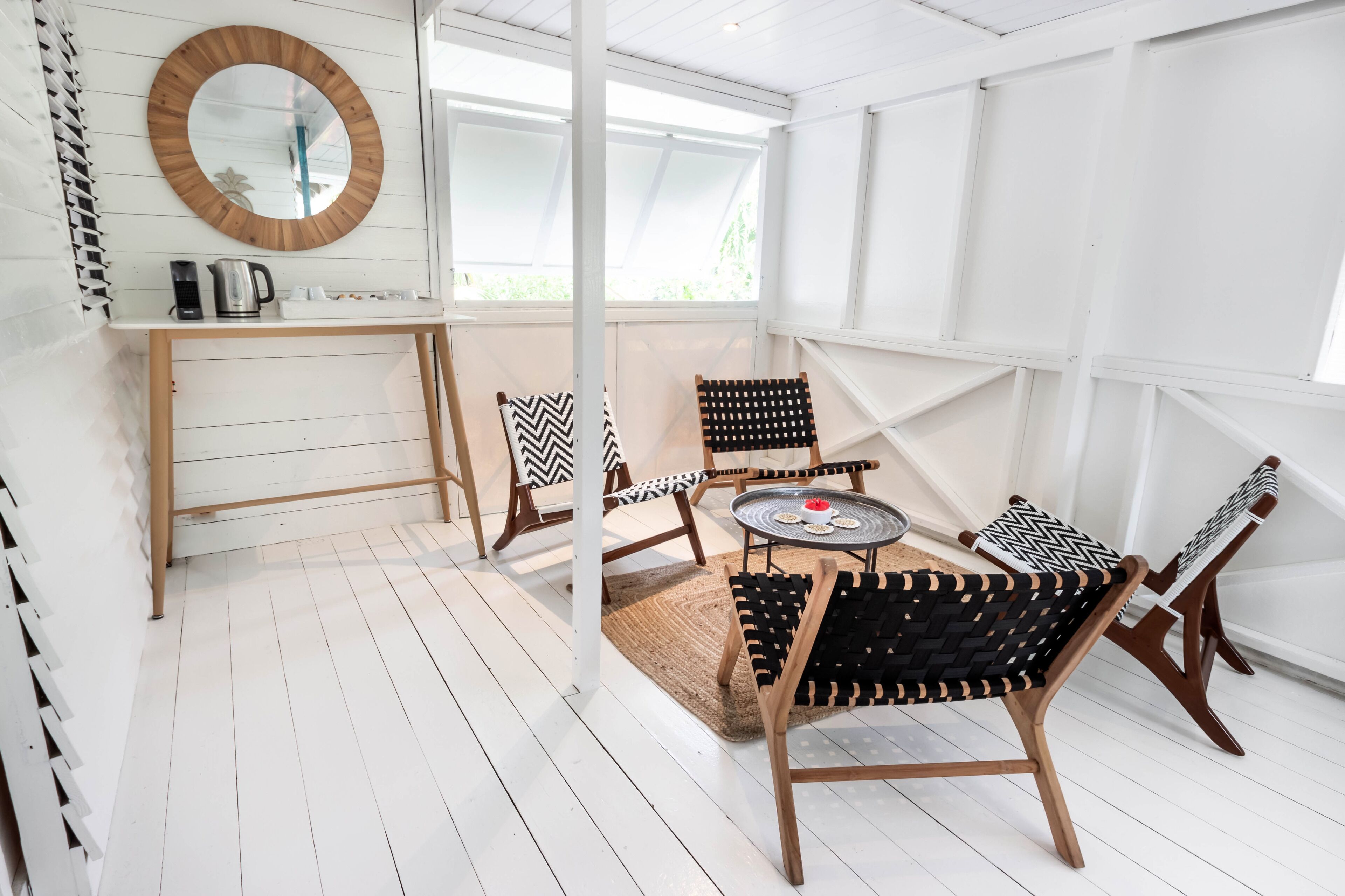 Una habitación luminosa y blanca con un espejo redondo, una cafetera y dos sillas con cojines de estampado blanco y negro.