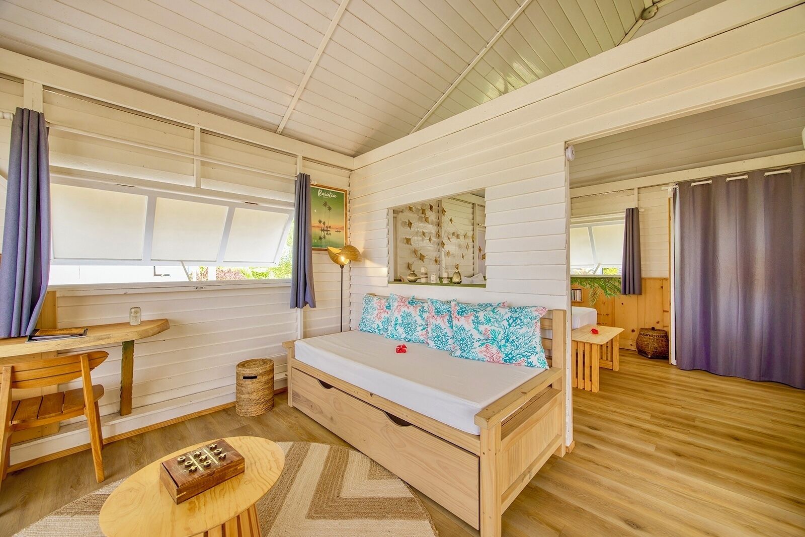 A cozy room with a wooden bed, a small table, and a chair. There is a window with curtains and a view of greenery outside.