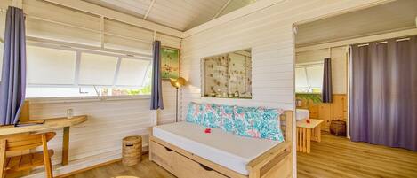 A cozy room with a wooden bed, a small table, and a chair. There is a window with curtains and a view of greenery outside.