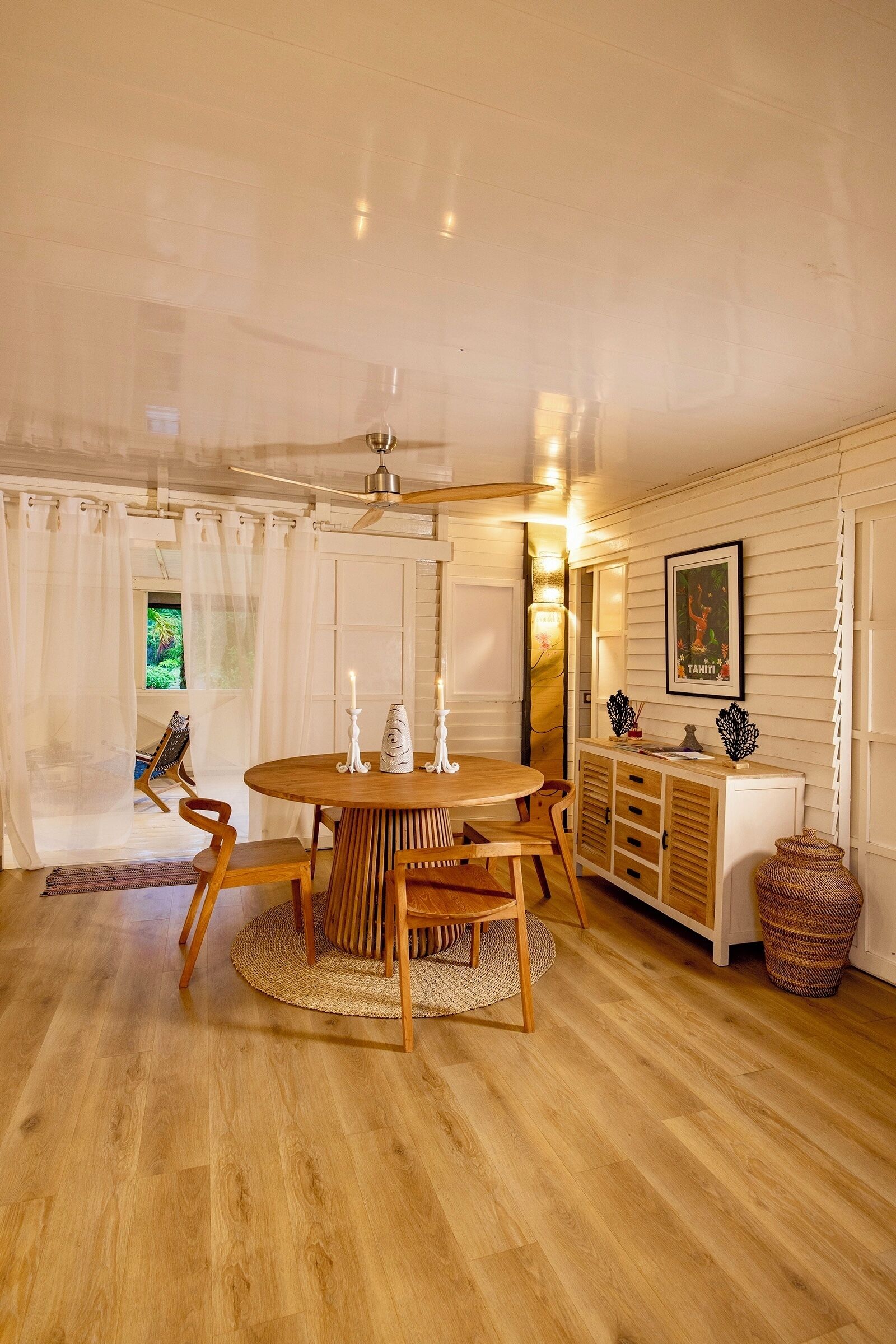Un coin repas avec une table et des chaises en bois, un ventilateur de plafond et une vue sur l’extérieur par une fenêtre.