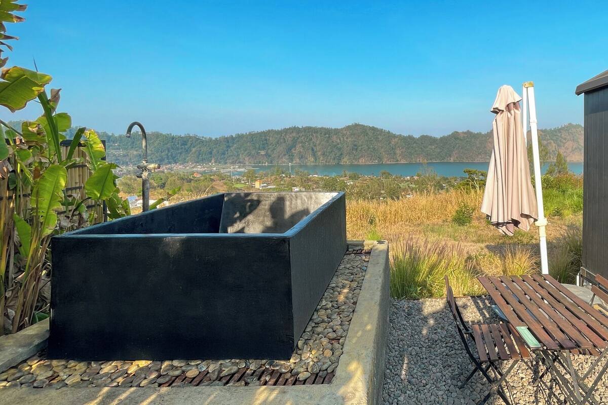 A rectangular outdoor bathtub with a stone border, a wooden bench, and a covered umbrella.