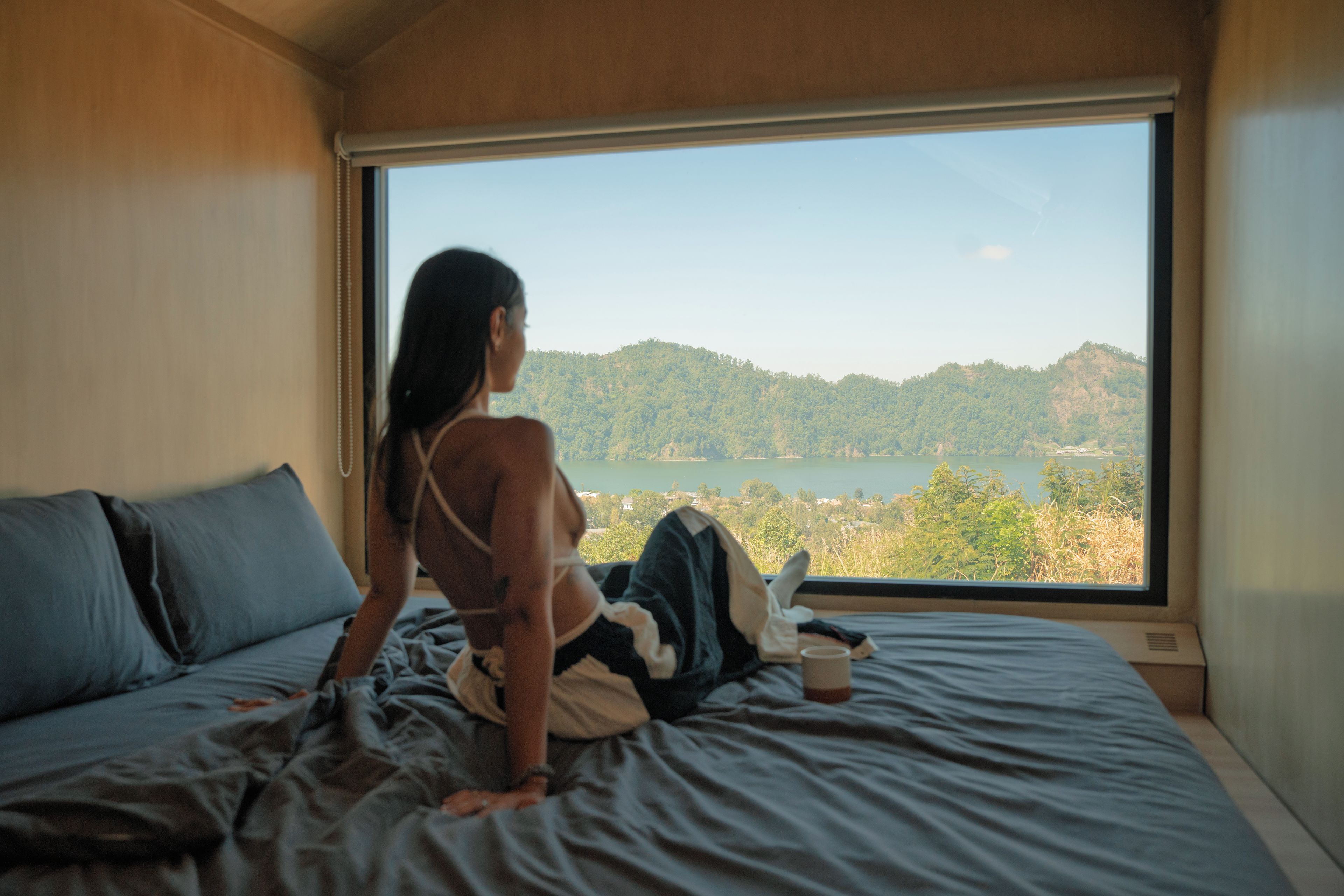 A person sitting on a bed with a window view of a lake and hills.