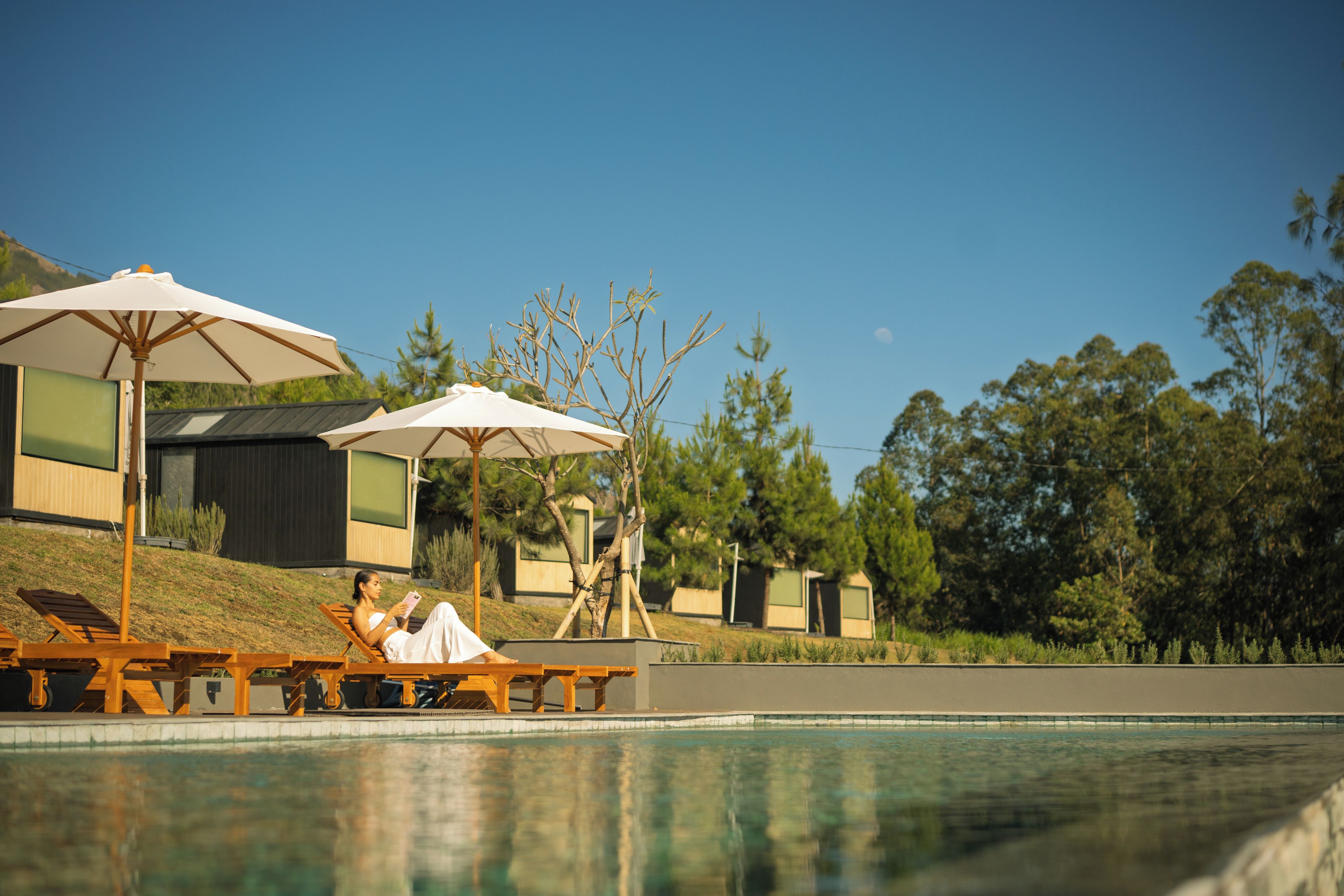 A poolside area with lounge chairs and umbrellas, a person reading, and a building with a modern design in the background.