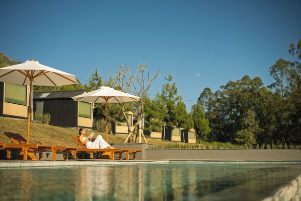 A poolside area with lounge chairs and umbrellas, a person reading, and a building with a modern design in the background. - Bobocabin Kintamani (Kintamani)
