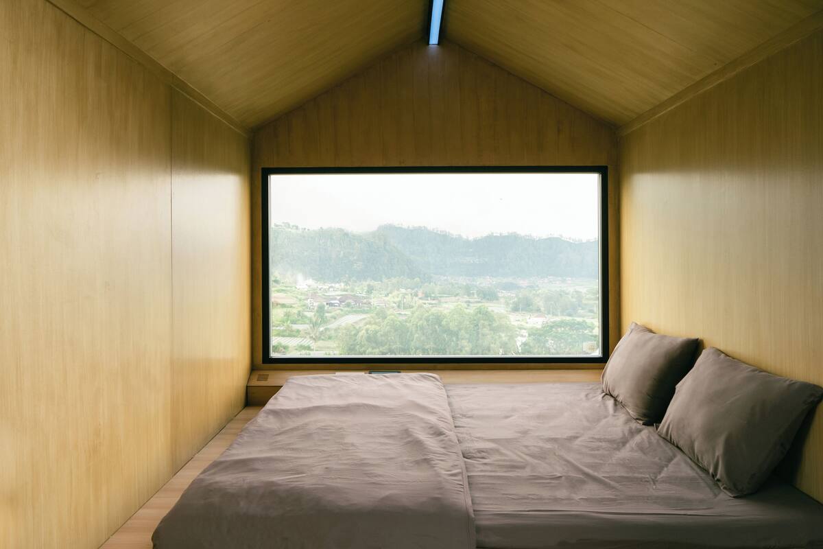 A bedroom with a large window, wooden walls, and a bed with pillows.