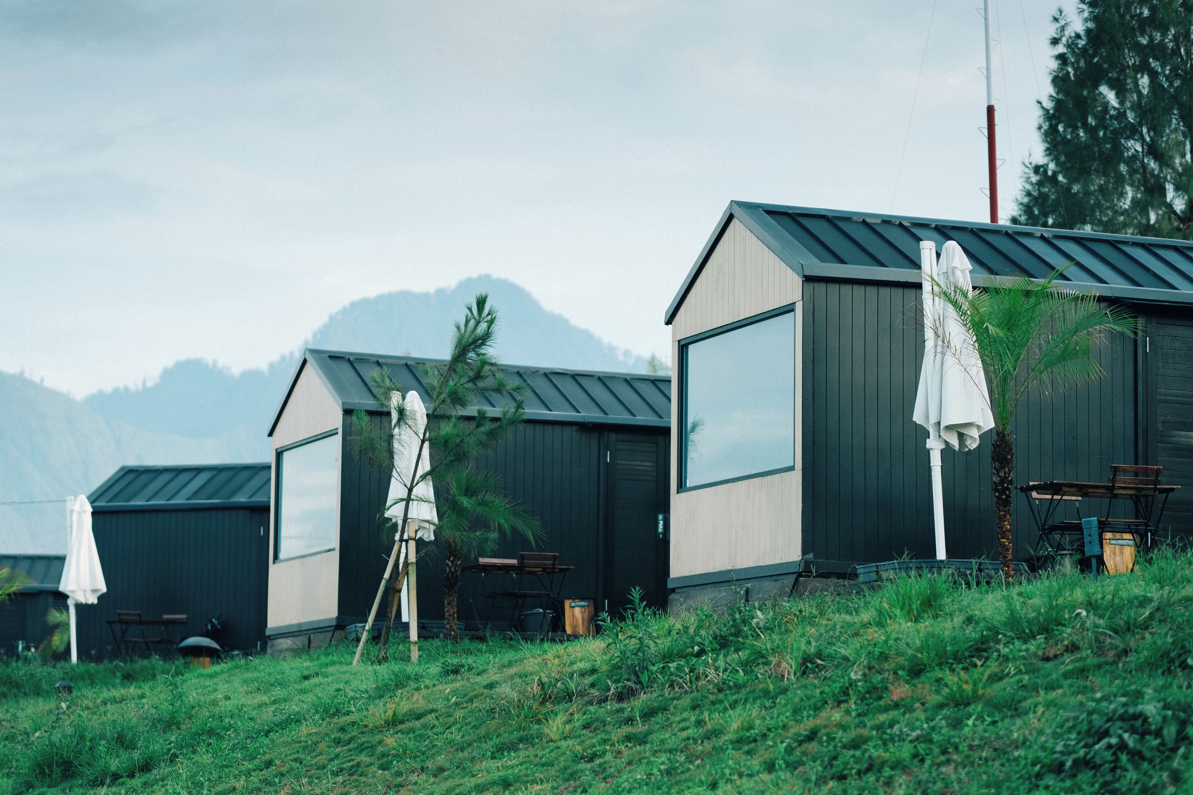 A row of modern cabins with large windows, outdoor seating, and a view of mountains.