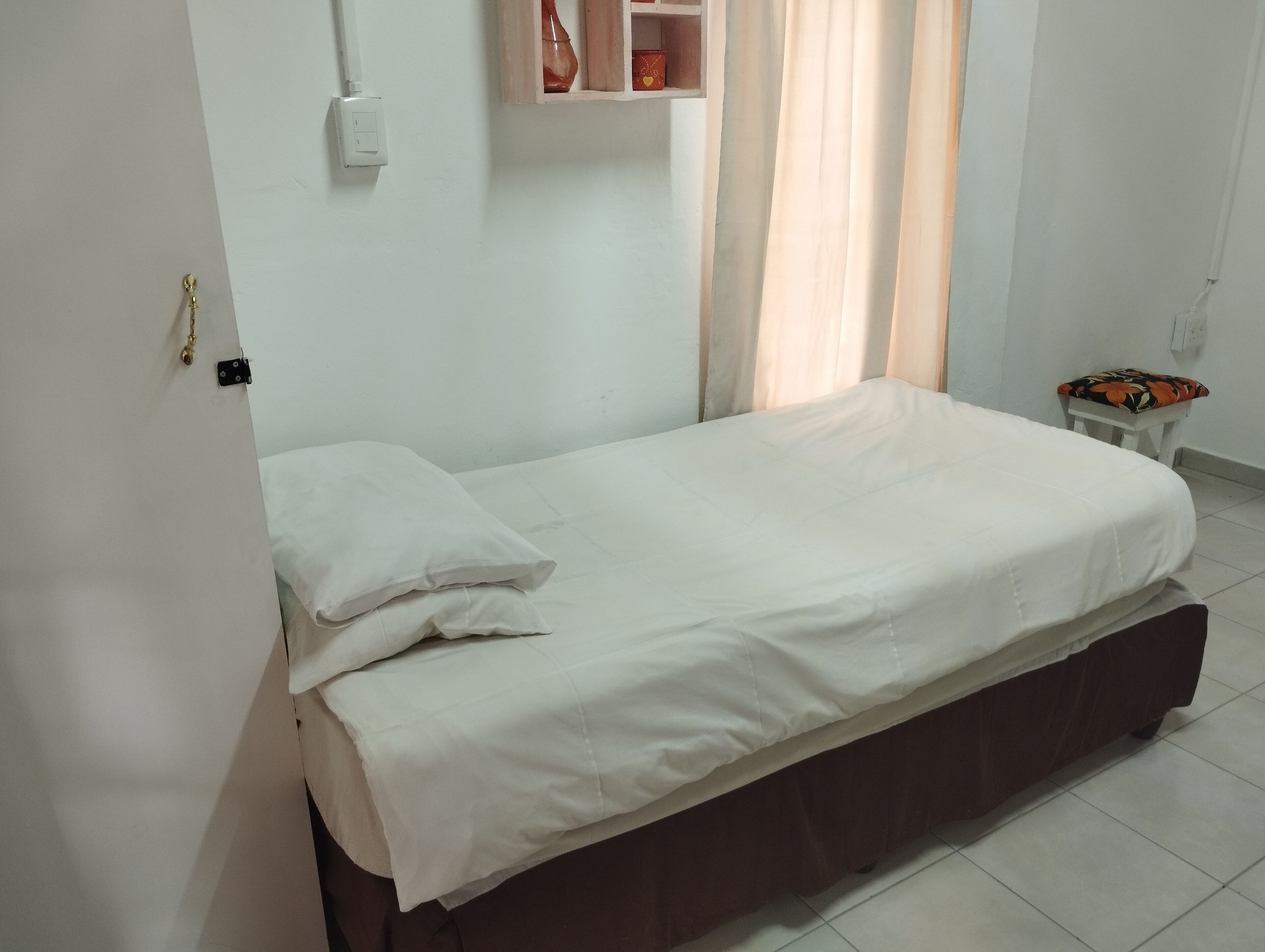 A single bed with a wooden frame and white bedding in a room with white walls, a small table with a patterned cushion, and a window with curtains.
