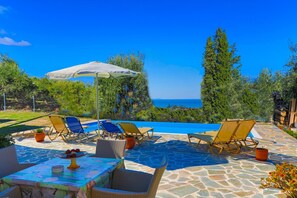 Zona piscina con poltrone prendisole, un tavolo con un vassoio di frutta e vista sul mare.