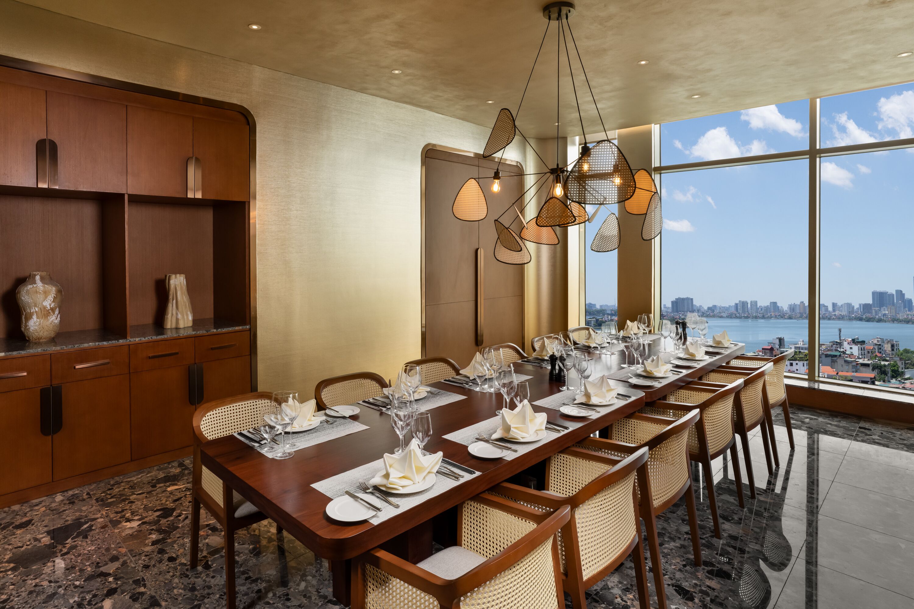 A dining area with a long table set for a meal, large windows offering a city view, and a modern chandelier.
