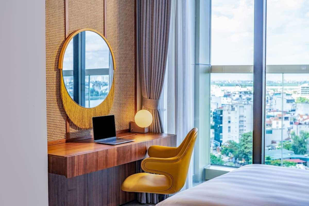 Habitación de hotel con un escritorio de madera, una laptop, una silla amarilla, un espejo redondo y un ventanal con vistas a la ciudad.