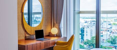 Habitación de hotel con un escritorio de madera, una laptop, una silla amarilla, un espejo redondo y un ventanal con vistas a la ciudad.