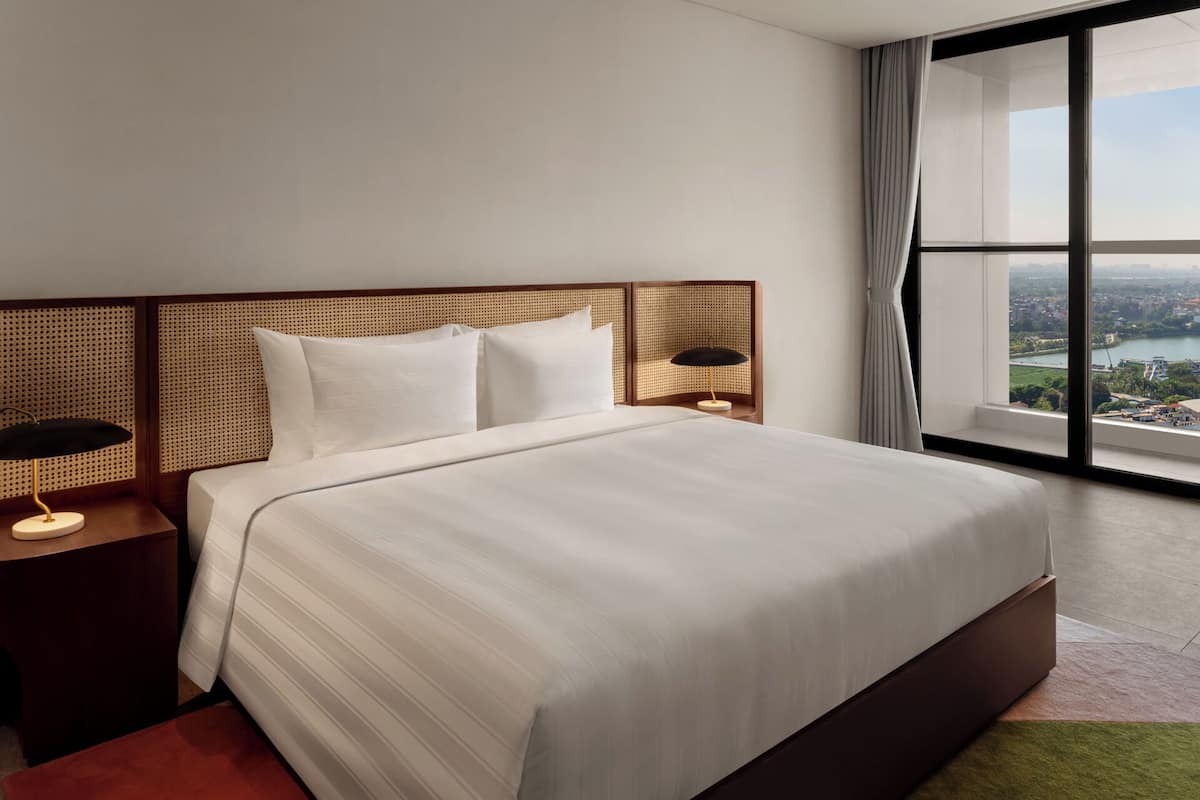 Habitación de hotel moderna con una cama grande, una mesita de noche con lámpara y vistas a un paisaje urbano a través de un ventanal amplio.