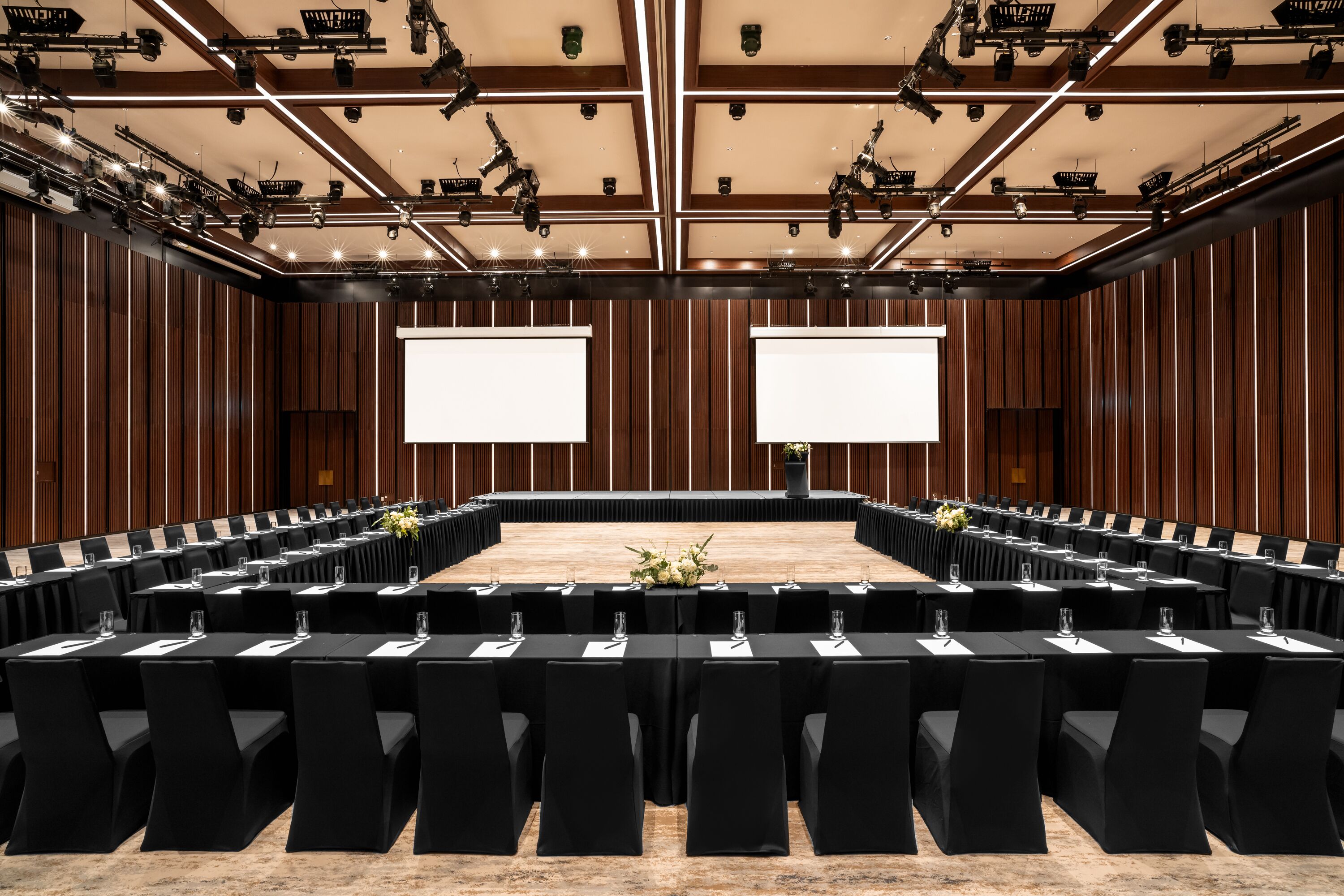 A conference room with a long table, rows of chairs, and two large screens.