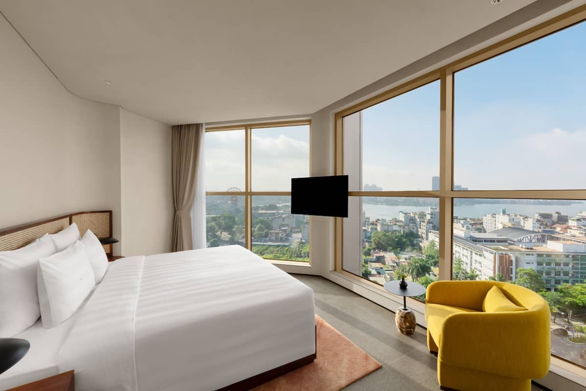 Una habitación de hotel moderna con una cama grande, un sillón amarillo y vistas a la ciudad a través de amplios ventanales.