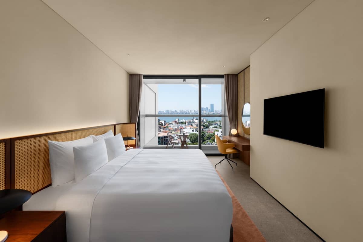 Habitación de hotel con una cama grande, un televisor de pantalla plana, un escritorio con silla y vistas a la ciudad a través de un amplio ventanal.
