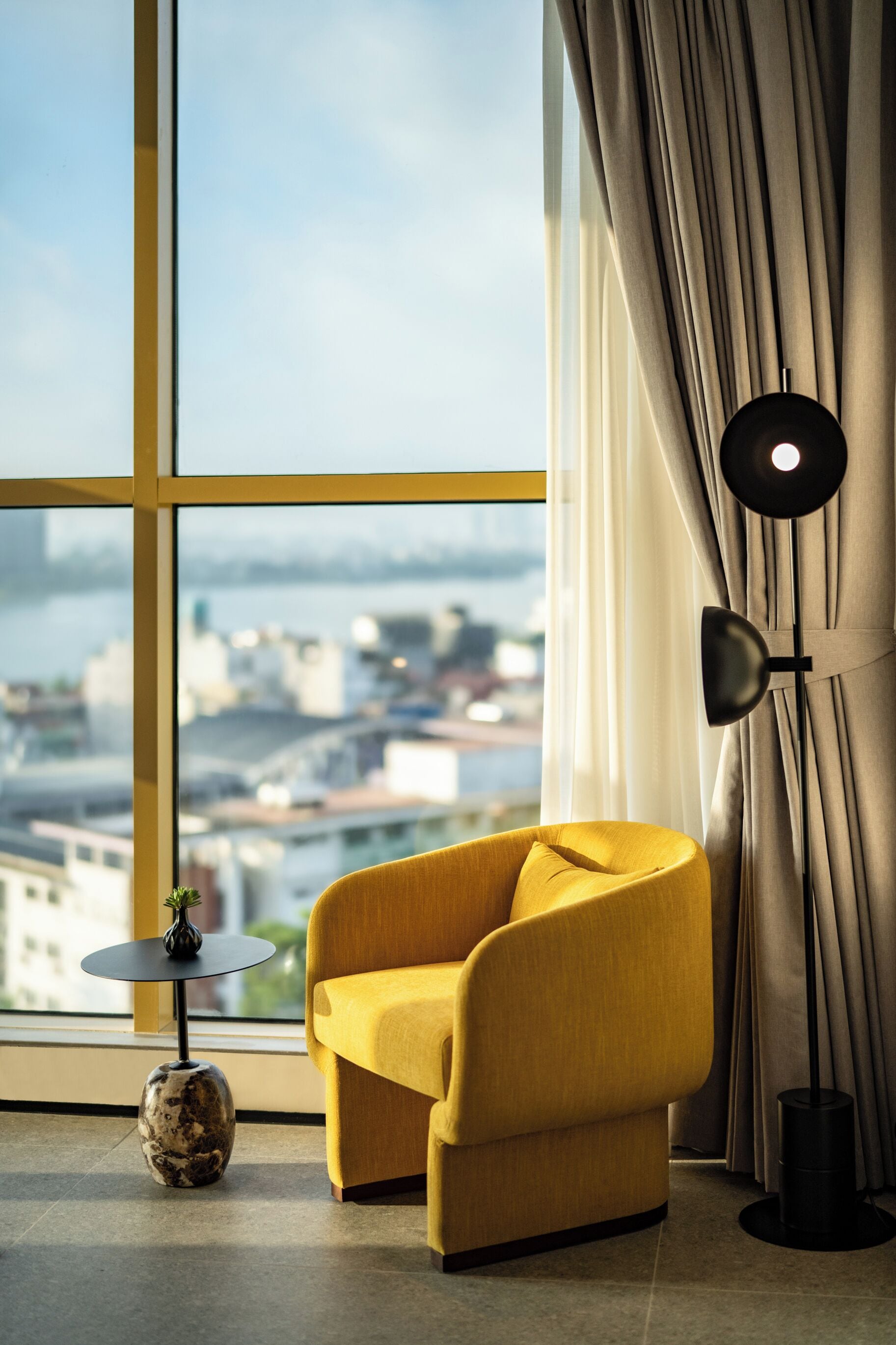 Een moderne kamer met een gele fauteuil, een ronde tafel met een vaas, en een vloerlamp. Een groot raam biedt uitzicht over de stad.