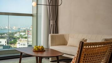 A modern living room with a sofa, a wooden coffee table with pears, a wicker chair, and a view of a cityscape through large windows.
