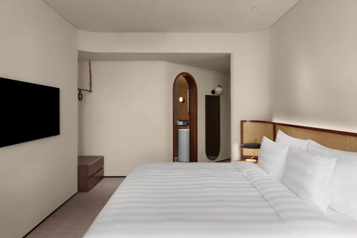 Una habitación de hotel con una cama grande, un televisor de pantalla plana montado en la pared, un cabecero de madera y una mesita de noche con una lámpara.