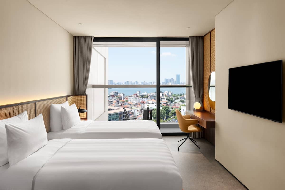 Habitación de hotel con dos camas, un escritorio con silla, televisor y un ventanal con vista a la ciudad.